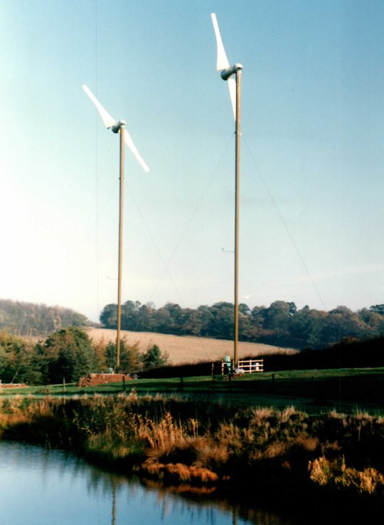 Two Carter 25 kW turbines on slender guyed towers above a pond and reeds at Beacon Farm