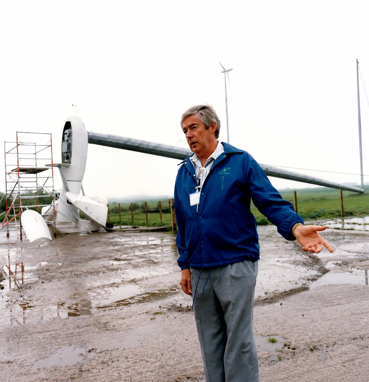 Tony Marmont in a blue jacket gesturing as he speaks, standing on the muddy ground at Great Orton with a Carter turbine tilted down behind him and another standing in the distance